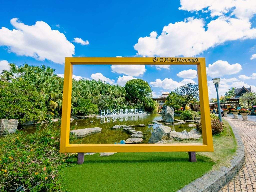 View of the entrance area in Riyuegu Hotsprings Park featuring a golden picture- frame installation against a backdrop of lush greenery and blue skies filled with clouds.